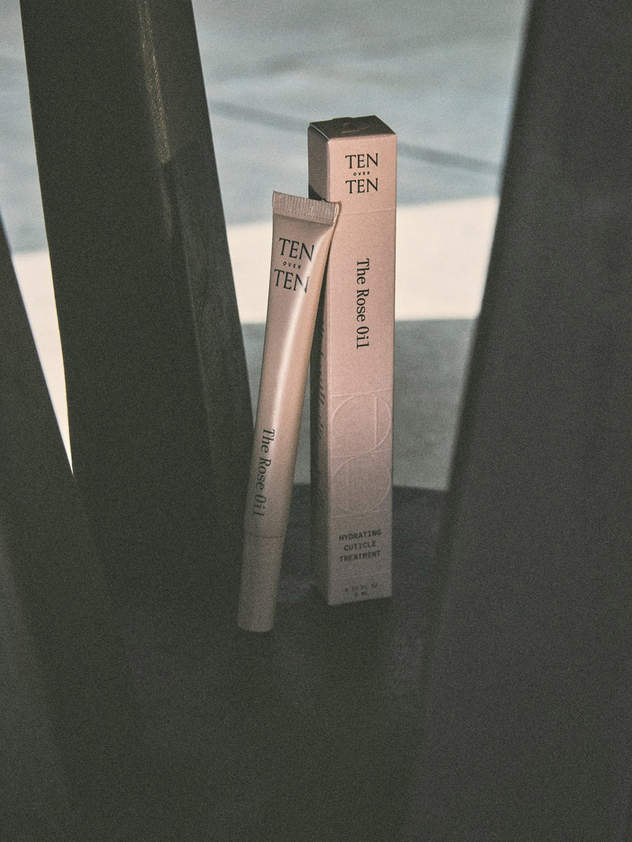 A tube and box of TEN OVER TEN - THE ROSE OIL CUTICLE TREATMENT stand upright on a dark, angular surface under soft lighting, emphasizing the brand’s hydrating formula for restoring nails and cuticles.