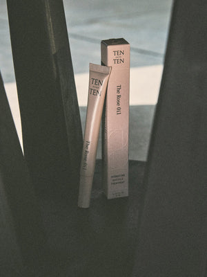 A tube and box of TEN OVER TEN - THE ROSE OIL CUTICLE TREATMENT stand upright on a dark, angular surface under soft lighting, emphasizing the brand’s hydrating formula for restoring nails and cuticles.