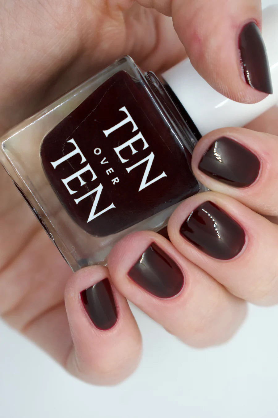 A glass bottle of TENOVERTEN - NAIL POLISH from the brand TEN OVER TEN, featuring a white cap and a dusty pink nail color. The front label displays "TEN OVER TEN" in white uppercase letters. This vegan, eight-free formula ensures both beauty and peace of mind. The background is plain and light-colored.
