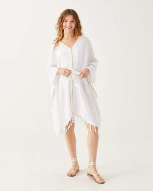 A woman with wavy hair smiles in the MERSEA - BEACH KAFTAN, a loose white Turkish cotton cover-up with fringe and gold trim, tied at the waist. She pairs it with sandals, showcasing MERSEA’s effortless resort style against a white background.