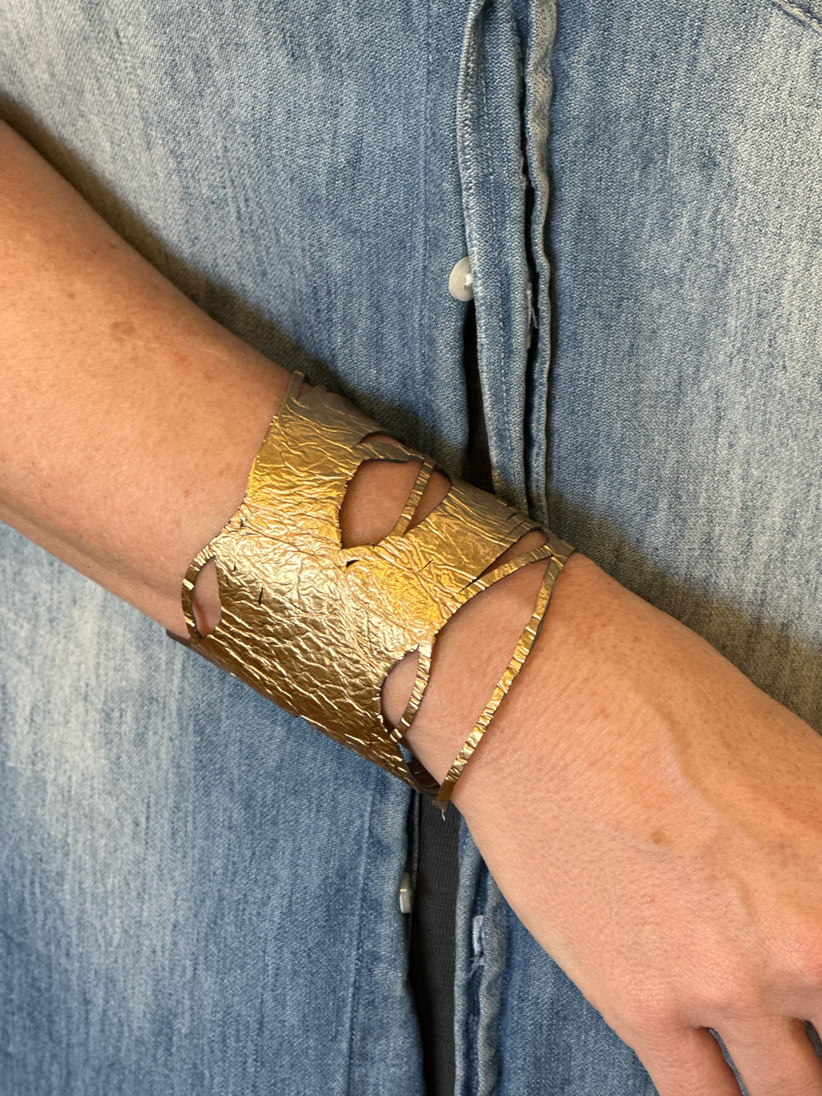 A person wearing the ILLUSIONISM BRACELET IN GOLD AND SILVER (REVERSIBLE) from SI SABATO ISABEL on their wrist. The handmade jewellery, crafted by an Argentinian artist, features irregular cut-out shapes and an intricate, hammered surface. The person is dressed in a faded blue denim shirt with white buttons.