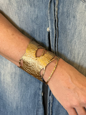 A person wearing the ILLUSIONISM BRACELET IN GOLD AND SILVER (REVERSIBLE) from SI SABATO ISABEL on their wrist. The handmade jewellery, crafted by an Argentinian artist, features irregular cut-out shapes and an intricate, hammered surface. The person is dressed in a faded blue denim shirt with white buttons.