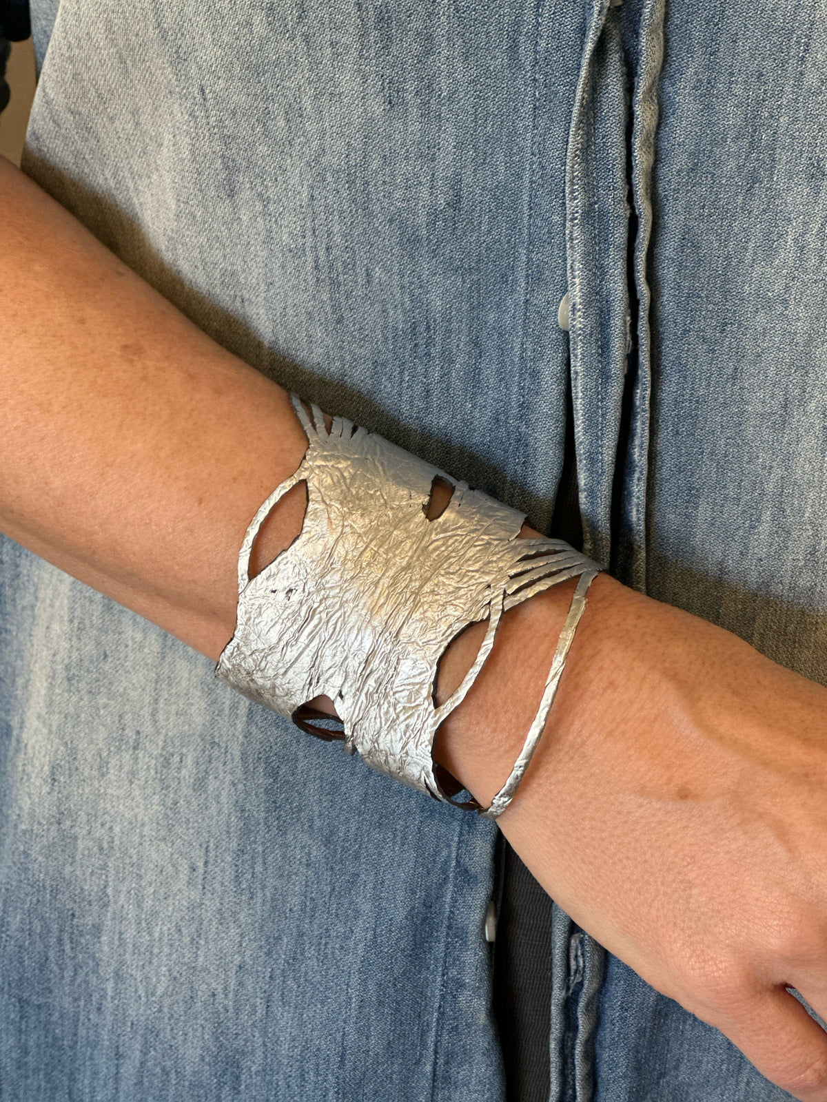 A person wearing the ILLUSIONISM BRACELET IN GOLD AND SILVER (REVERSIBLE) from SI SABATO ISABEL on their wrist. The handmade jewellery, crafted by an Argentinian artist, features irregular cut-out shapes and an intricate, hammered surface. The person is dressed in a faded blue denim shirt with white buttons.
