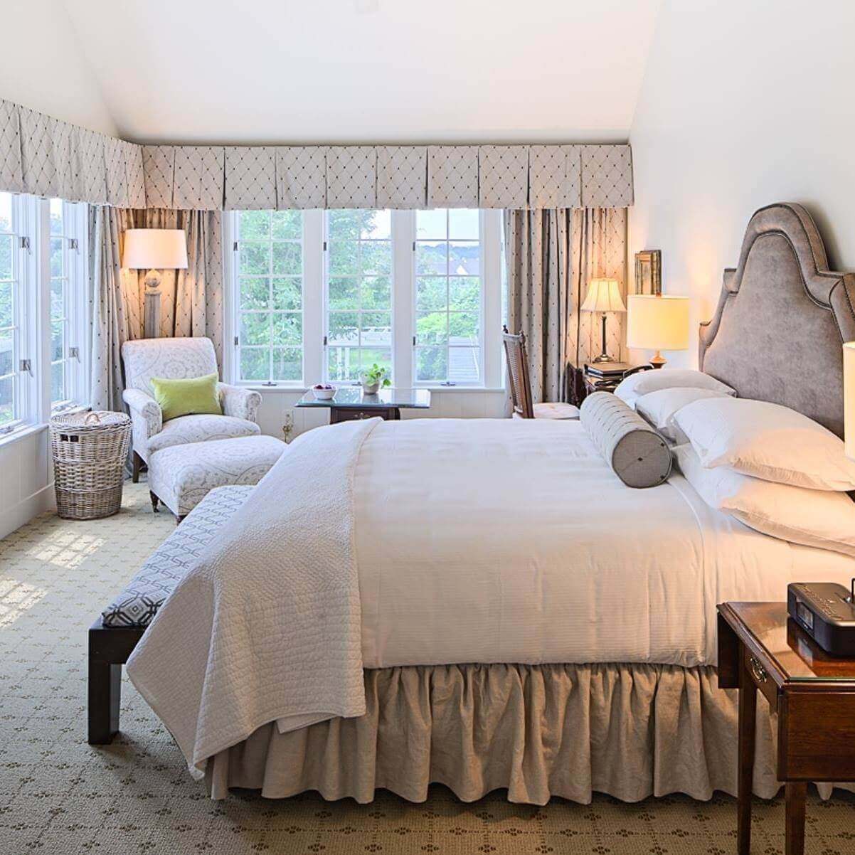 A cozy, well-lit bedroom featuring a large bed with white bedding, a padded headboard, and a beige skirt. Large windows with patterned curtains line two walls, letting in natural light. There's also a cushioned lounge chair, a basket, a side table, and modern lamps. Fearrington Village