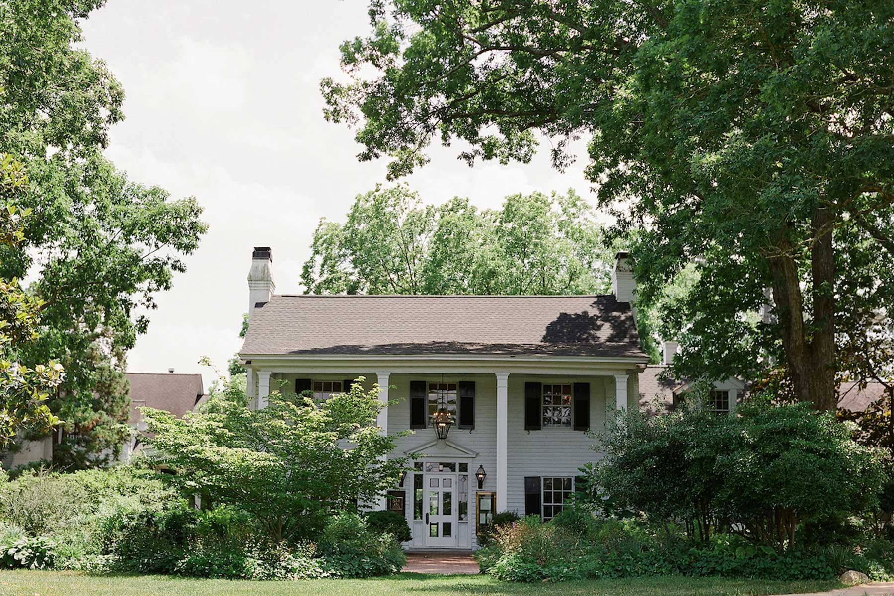 A charming white two-story house sits amidst lush greenery and tall trees. The house has a gabled roof, black shutters, a chimney on each side of the roof, and a covered front porch with an arched entryway. The scene is serene and bathed in natural daylight. Fearrington Village