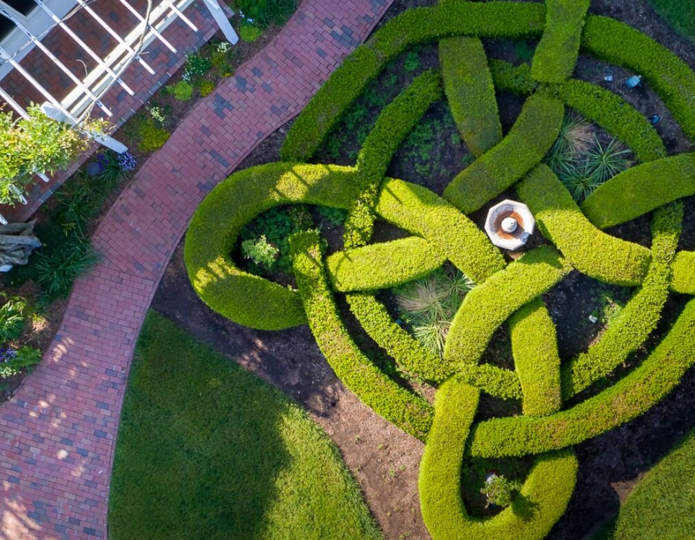 Aerial view of a garden with neatly trimmed, interwoven hedges forming an intricate knot pattern. A person in a wide-brimmed hat is standing in the center of the design. Surrounding the hedge, a brick pathway runs alongside a lawn and part of a building is visible. Fearrington Village