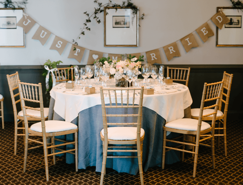 A decorated round table set for a wedding reception, surrounded by wooden chairs. The table is adorned with a white and blue tablecloth, floral centerpiece, and glassware. A "Just Married" banner hangs on the wall behind the table, with framed pictures above. Fearrington Village