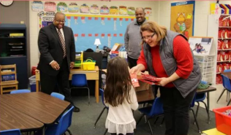 A smiling teacher in a red shirt and gray vest hands a wrapped gift to a young girl in a classroom. Two men, one in a suit and another in casual attire, look on with smiles. The room is decorated with colorful artwork and educational materials. Fearrington Village