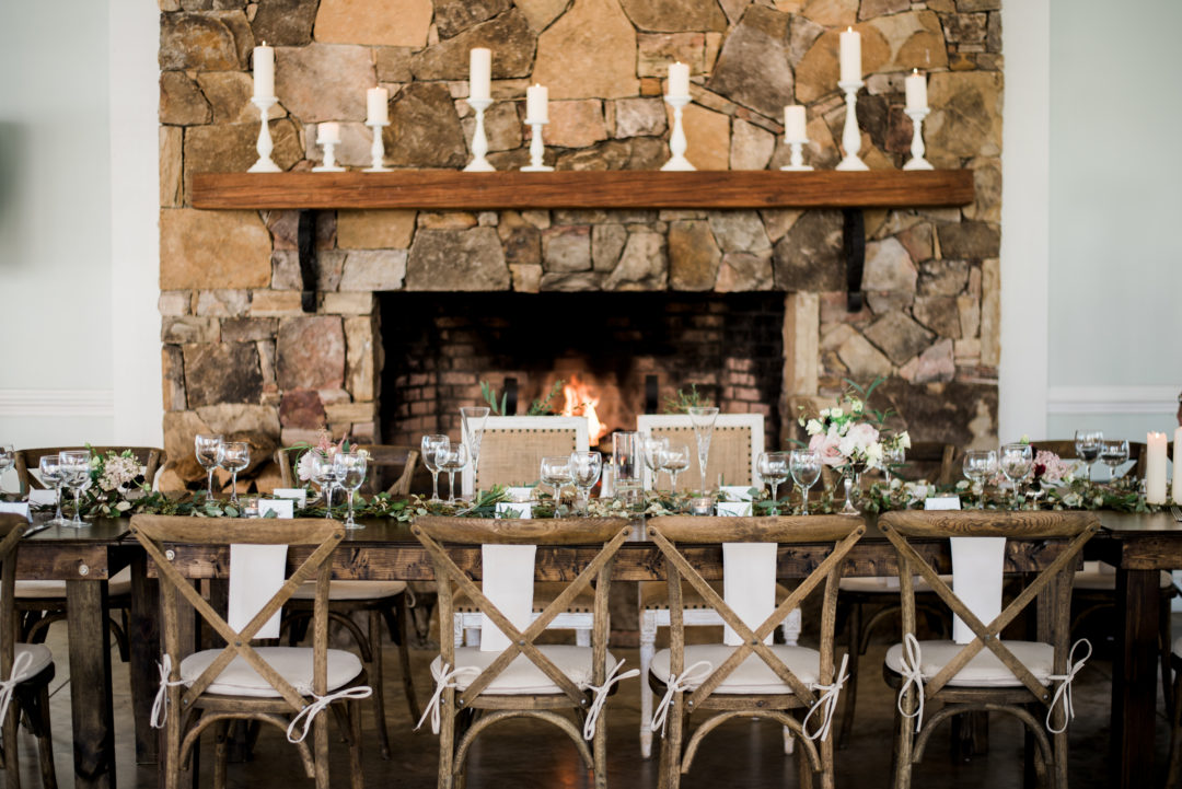 A rustic dining setup with a wooden table adorned with greenery, candles, and elegant place settings in front of a stone fireplace. Wooden cross-back chairs, some with white ribbons, enhance the cozy ambiance. The fireplace is lit, creating a warm atmosphere. Fearrington Village