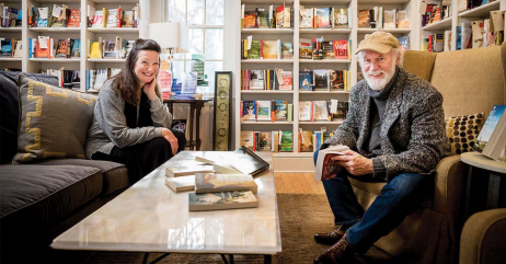 Two people are seated in a cozy bookstore. A woman with long dark hair and a gray sweater sits on a sofa with her hand on her face, smiling. A bearded man in a hat and gray jacket sits in an armchair, holding an open book and smiling. Bookshelves are in the background. Fearrington Village