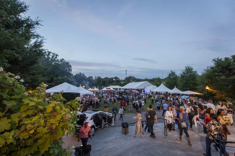 A lively outdoor event with numerous people gathered under white tents. Attendees are strolling, socializing, and enjoying the evening. Surrounding trees and a clear sky frame the scene. A few cars and a mix of casual and formal attire can be seen. Fearrington Village