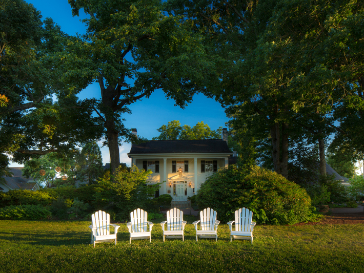 A quaint white house with dark shutters is surrounded by lush greenery and tall trees. On the well-manicured lawn in front, five white Adirondack chairs are neatly arranged in a row, facing the house. The scene is peaceful with a clear blue sky. Fearrington Village