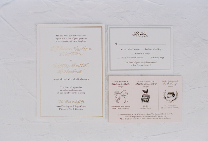 A wedding invitation suite includes a main invite with elegant gold font, an RSVP card, and two smaller inserts with details about pre-wedding events. The elements are laid out on a textured white surface. Each card has a classic and sophisticated design. Fearrington Village