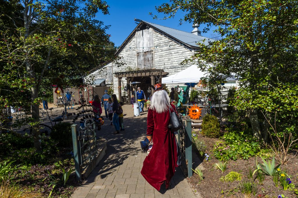 A person with long white hair wearing a red coat walks down a path toward a rustic barn. The setting is a lively outdoor market or event with various booths, decorations, and people milling around. Greenery surrounds the area, and the sky is clear and blue. Fearrington Village