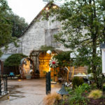 A quaint outdoor area with rustic charm features a weathered barn as the backdrop. Decorative items and lights adorn the space, while a sign in the foreground displays a poster about "Farmington Folk Art" from 2021. Lush greenery adds to the inviting ambiance. Fearrington Village