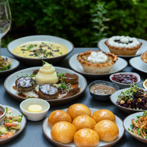 A table set with a variety of dishes including shrimp cocktail, bread rolls, mashed potatoes, meat with gravy, mixed vegetables, dessert pies, soups, and side sauces. Two empty wine glasses are placed on the table. The backdrop features greenery. Fearrington Village