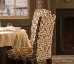 A cozy dining area featuring chairs with a geometric diamond pattern, a table with a white tablecloth set for a meal, and a lit fireplace in the background. The ambiance is warm and inviting, creating a perfect setting for a comfortable dining experience. Fearrington Village