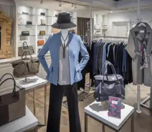 A clothing store interior featuring a mannequin dressed in a light blue blazer, white top, and black pants, with a black hat. Surrounding the mannequin are various handbags on tables, a scarf on a rack, and shelves with neatly folded clothing in the background. Fearrington Village