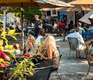 People dining and conversing at an outdoor restaurant with several tables. Umbrellas provide shade, and there are plants around. A variety of individuals are seated, enjoying food and drinks in a sunlit, relaxed atmosphere. Fearrington Village