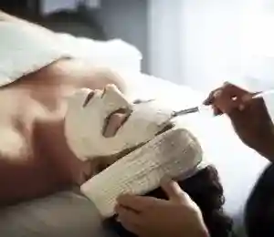 A person is lying down with a white facial mask applied to their face. Hands of another person are gently applying the mask with a brush. The individual receiving the treatment has a white headband to keep their hair back. The background is softly blurred. Fearrington Village