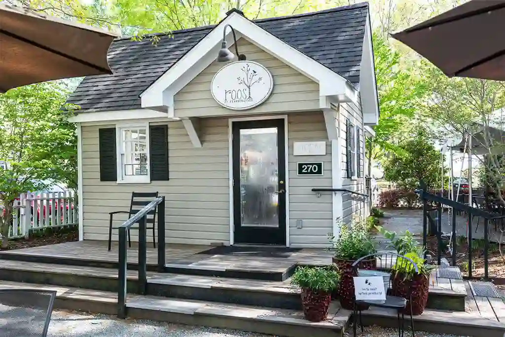 A small cottage-style building with beige siding and dark shutters. A sign above the door reads "The Roost." The entrance has a ramp and steps leading to it, with potted plants placed around the area. A black sign on the railing reads "Please do not climb ramp. Fearrington Village