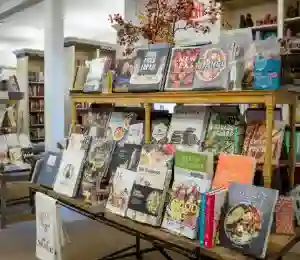 A library display features various cookbooks on wooden shelves. Book titles are visible and showcase a range of culinary topics. A branch with fall leaves decorates the top shelf. The rest of the library with more bookshelves can be seen in the background. Fearrington Village