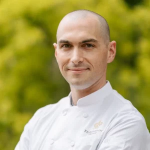 Paul Gagne. A man with a shaved head, wearing a white chef's coat, stands outdoors in front of a blurred background of yellow-green foliage. He has a slight smile and confident demeanor, and the uniform features an emblem on the upper left side. Fearrington Village