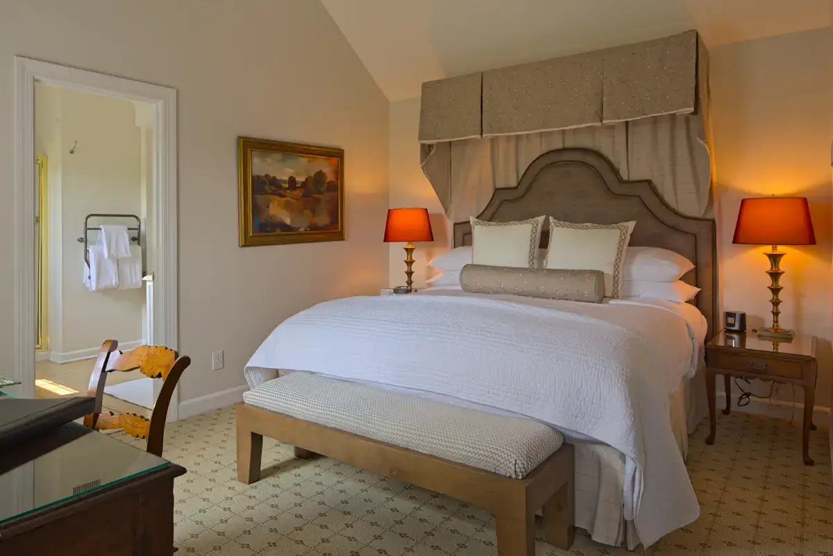 A cozy bedroom with a large, cushioned bed covered in white and beige linens. The bed has a decorative beige headboard and canopy. Two wooden nightstands, each with a red lamp, stand on either side. There's a bench at the foot of the bed and a painting on the wall. Fearrington Village