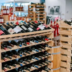 Free Wine Tasting - Drink Local 7 A wine store with multiple wooden racks filled with an assortment of wine bottles. The bottles are displayed horizontally in rows, with labels in various colors. In the background, additional wine racks and displays can be seen. Fearrington Village
