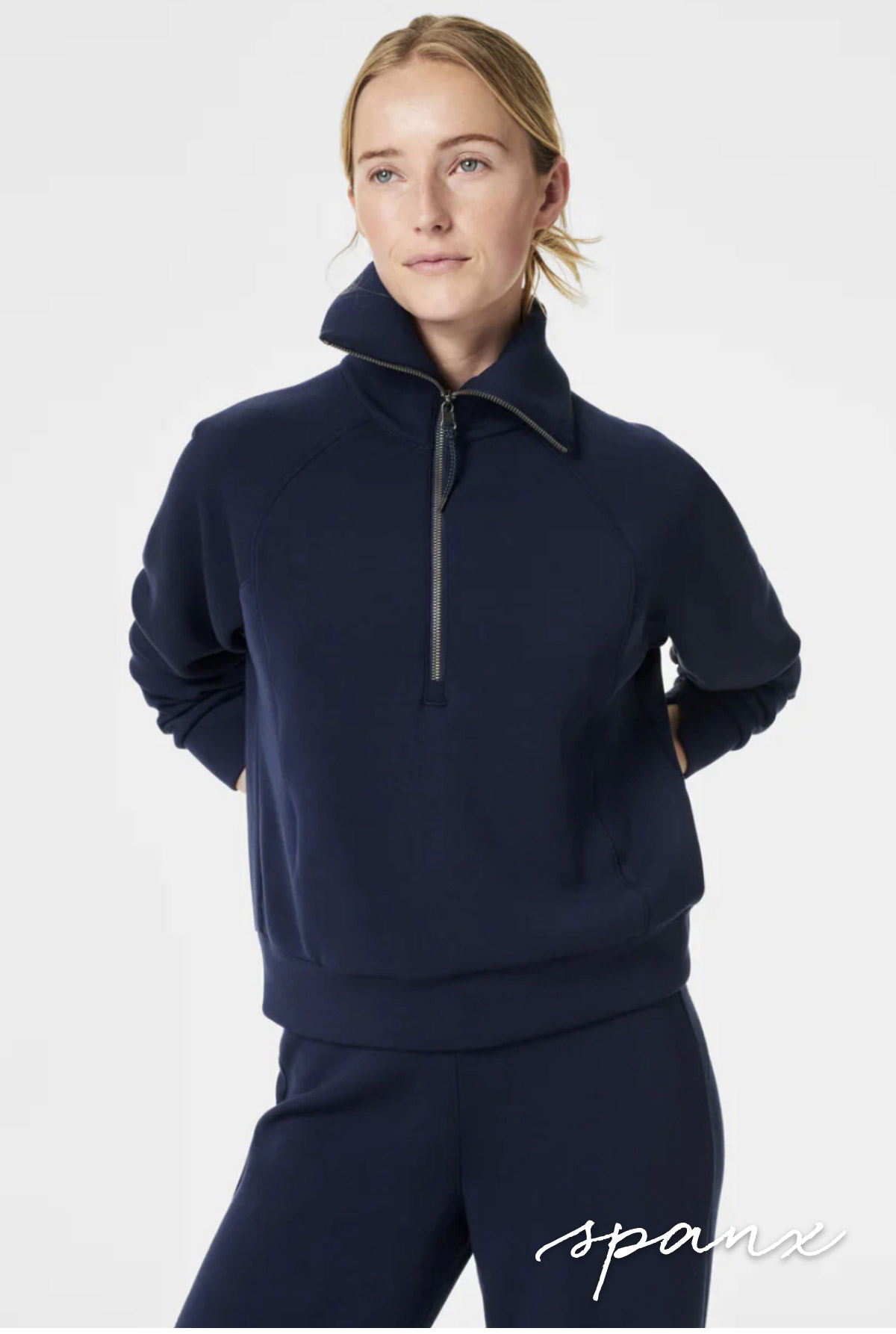 A person with blond hair stands wearing a navy blue long-sleeve, zip-up pullover with hands behind their back. The background is plain white, and the word "spanx" is written in cursive in the bottom right corner of the image. Fearrington Village