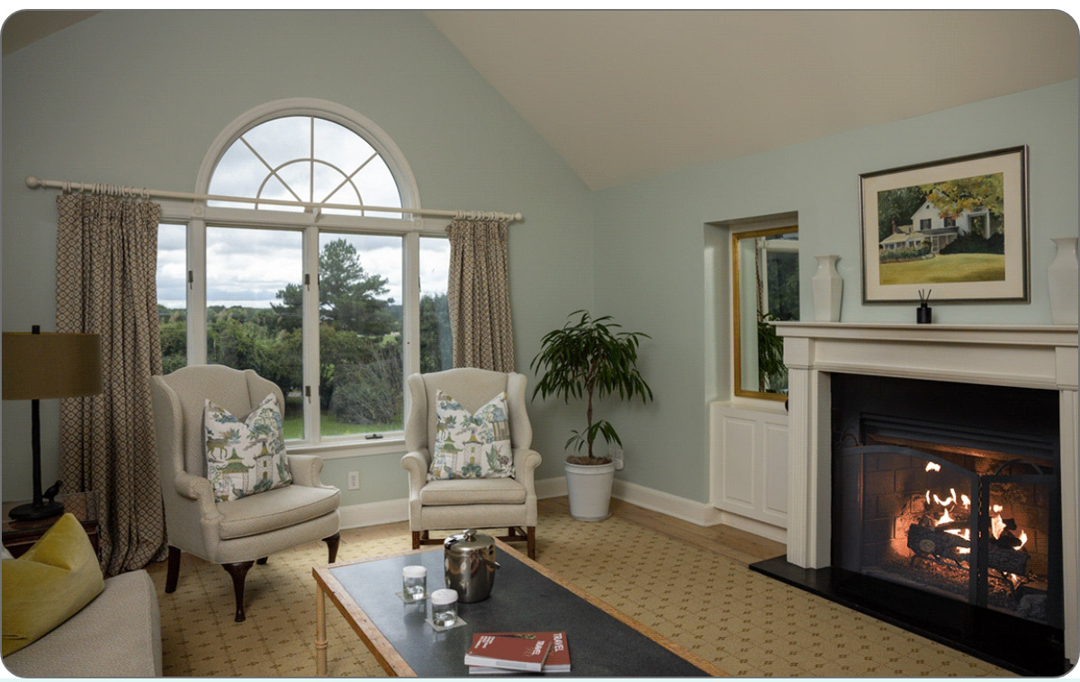 A cozy living room with a fireplace on the right, two armchairs with patterned cushions, and a large arched window showcasing an outdoor view. A potted plant is beside the fireplace, and a coffee table with books is in the foreground. Fearrington Village