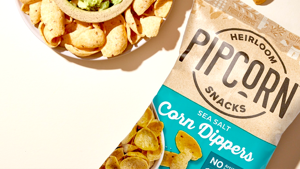 A bag of Pipcorn Heirloom Snacks labeled "Sea Salt Corn Dippers" next to a plate of corn chips with a green dip. The bag and plate are on a light-colored surface, creating a bright and clean presentation of the snack. Fearrington Village