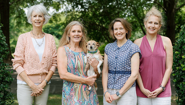 Four women stand together outside in a garden setting with trees in the background. One woman on the left wears a pink cardigan, another in a patterned dress holds a small dog, the third wears a blue blouse and light shorts, and the fourth wears a sleeveless pink top. Fearrington Village