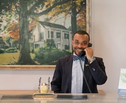 A man wearing a suit and bow tie stands behind a reception desk, smiling and talking on a phone. Behind him is a large framed painting of a house surrounded by trees. There are some items and brochures on the desk in front of him. Fearrington Village