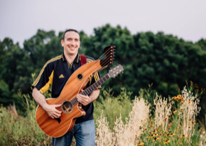 A man is standing in a field holding an acoustic harp guitar. He is smiling and wearing a dark sports jersey with yellow accents and jeans. The background has green trees and tall grass, with some flowers in the foreground. Fearrington Village