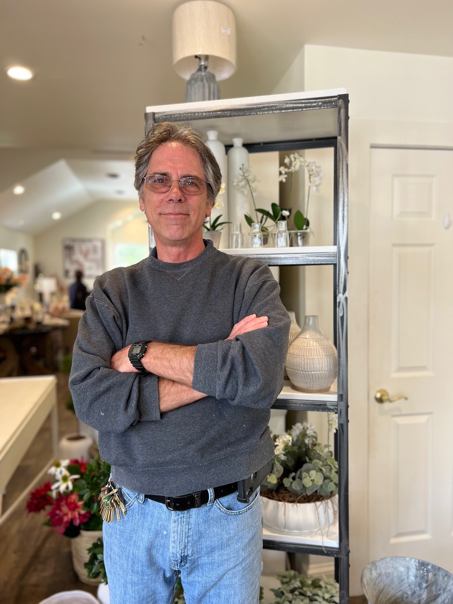 A man with gray hair and glasses stands confidently with arms crossed in a cozy indoor setting. He wears a gray sweatshirt and blue jeans, and is surrounded by potted plants and home decor items on shelves in the background. Fearrington Village