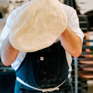 A person dressed in a white shirt and black apron is seen in a kitchen, stretching and tossing a large piece of pizza dough into the air. Shelves with baking trays and a blurry background suggest a busy kitchen environment. Fearrington Village