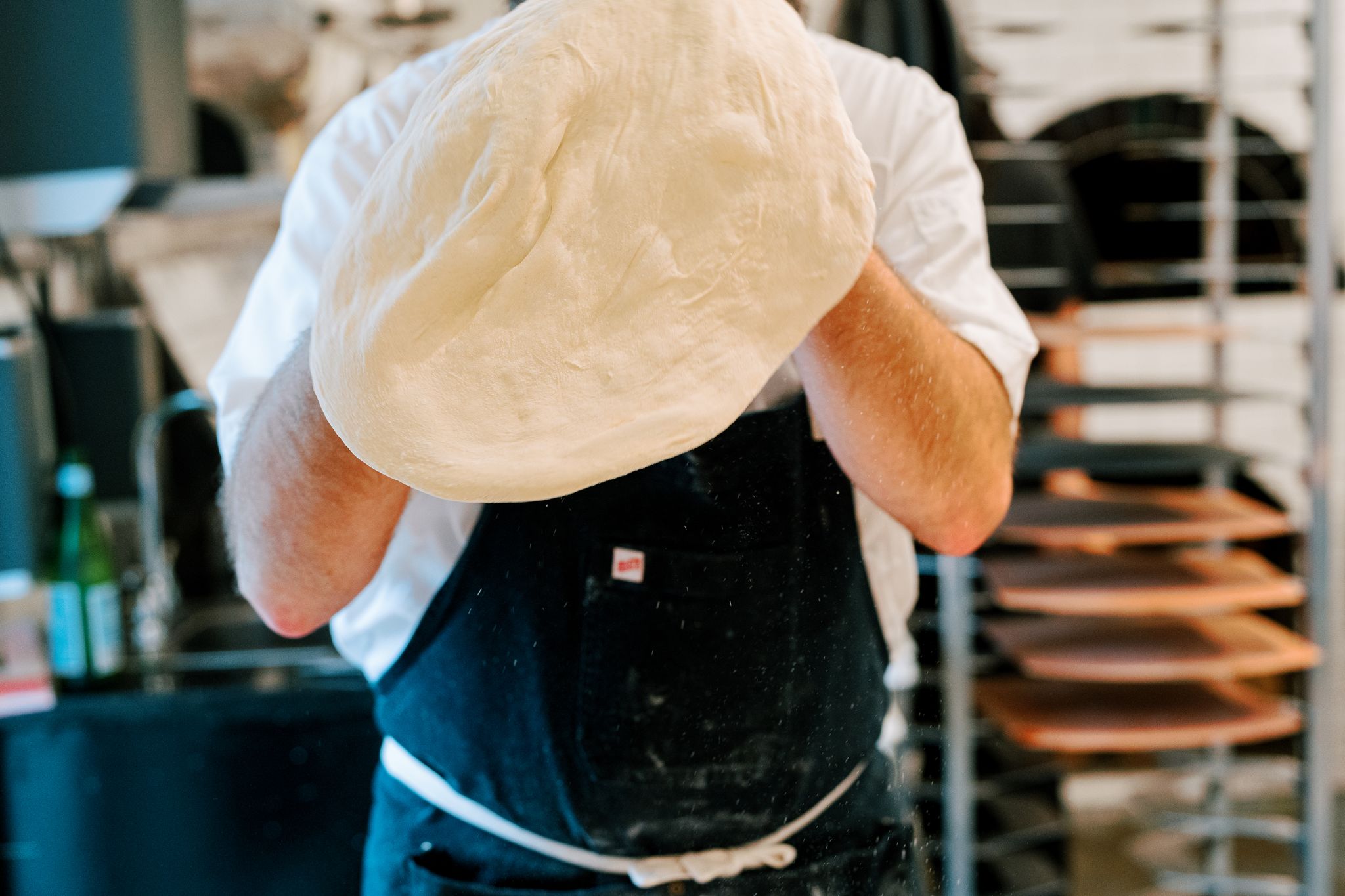 A person dressed in a white shirt and black apron is seen in a kitchen, stretching and tossing a large piece of pizza dough into the air. Shelves with baking trays and a blurry background suggest a busy kitchen environment. Fearrington Village