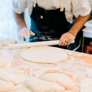 A person in a white shirt and black apron rolls out dough on a floured wooden surface. Several pieces of dough are laid out on the table, with kitchen equipment visible in the background. Fearrington Village