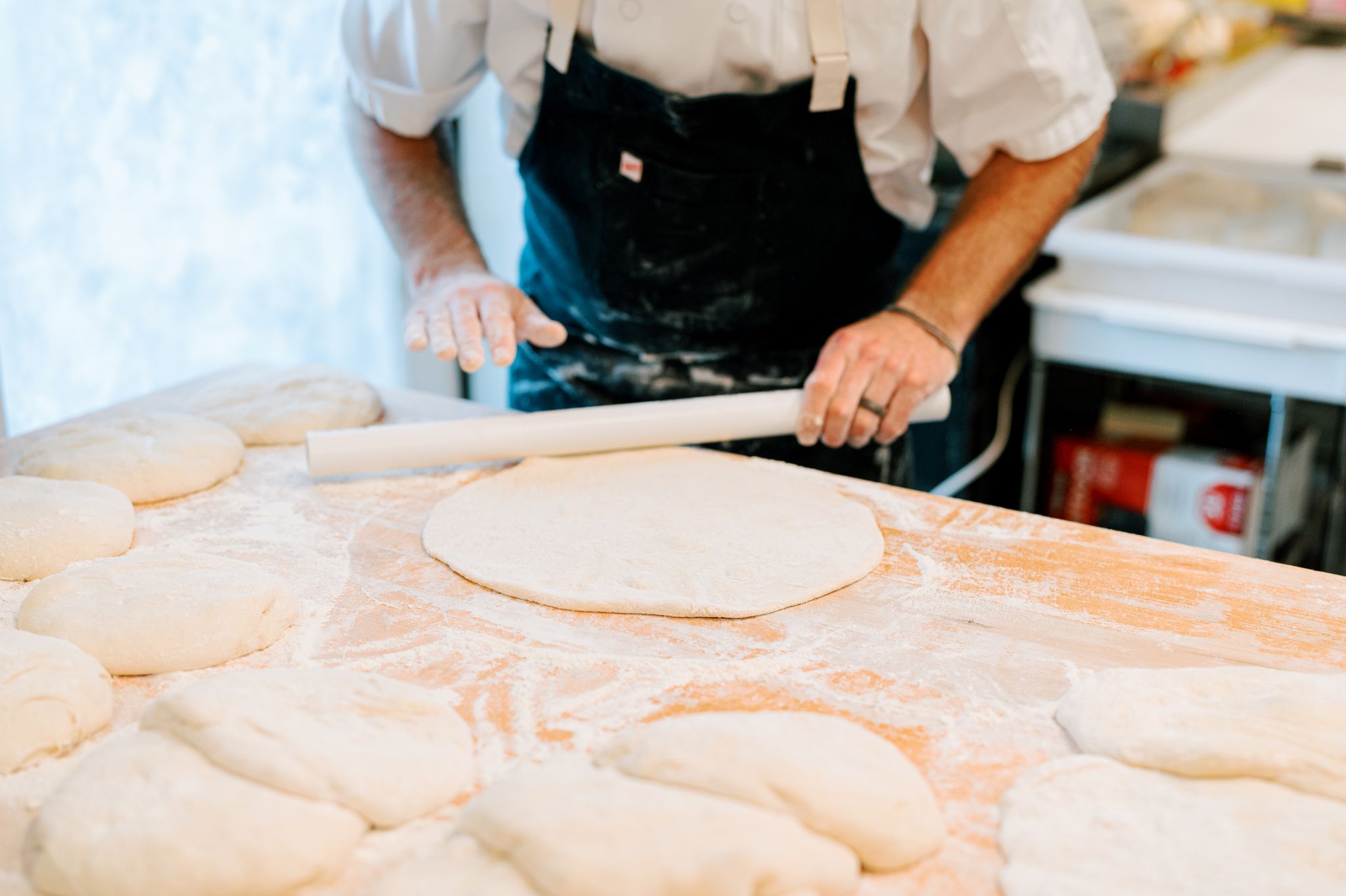 A person in a white shirt and black apron rolls out dough on a floured wooden surface. Several pieces of dough are laid out on the table, with kitchen equipment visible in the background. Fearrington Village