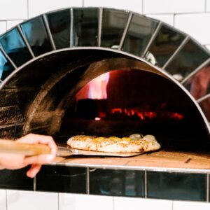 A hand using a pizza peel to place a pizza into a traditional wood-fired oven with a visible flame. The pizza crust is golden brown and bubbly. The oven is made of black tiles with a white-tiled wall in the background. Fearrington Village