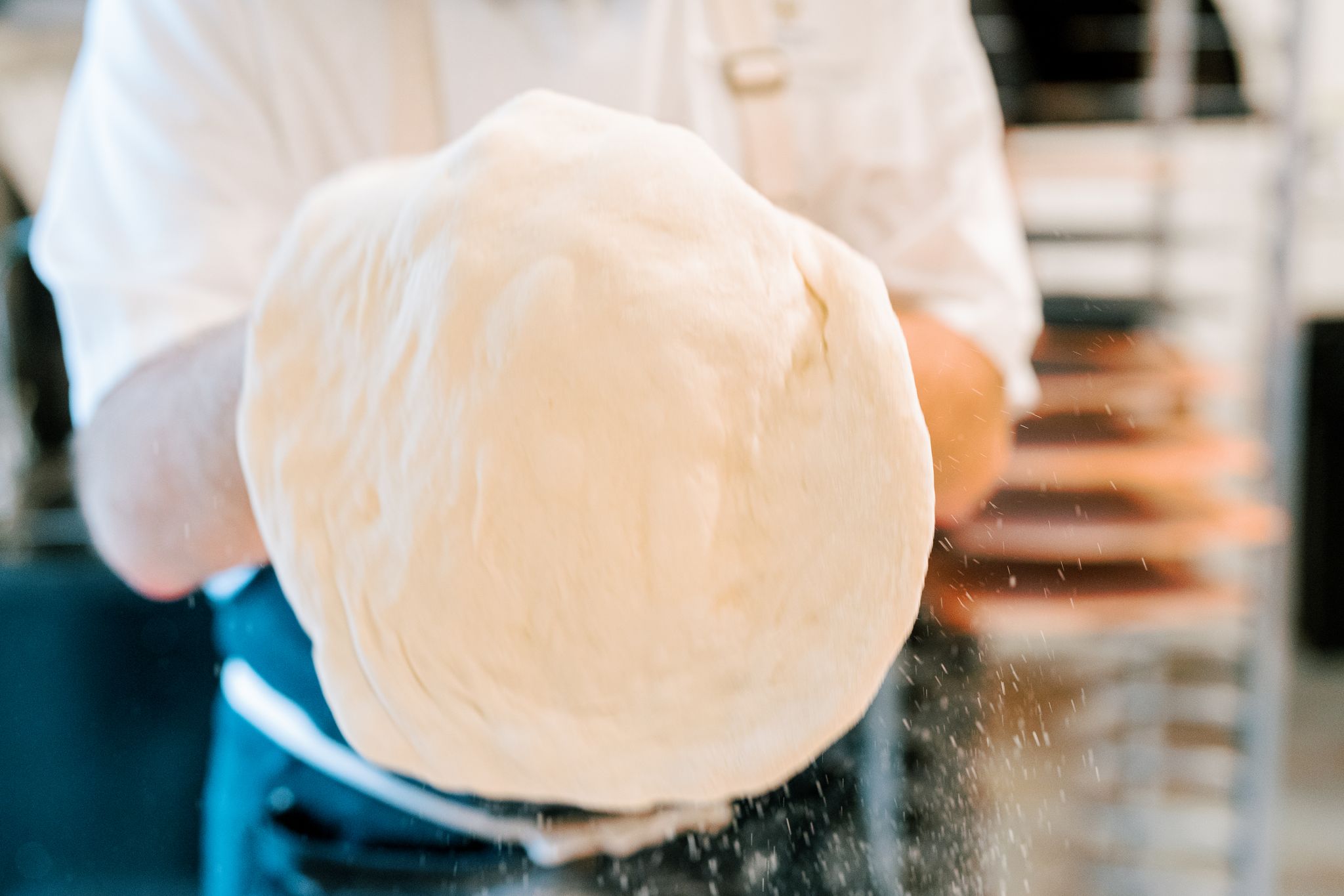 A person wearing a white shirt and apron is holding up a large round piece of pizza dough with both hands. The dough is being stretched and appears slightly airborne with motion blur. The background shows a kitchen environment. Fearrington Village