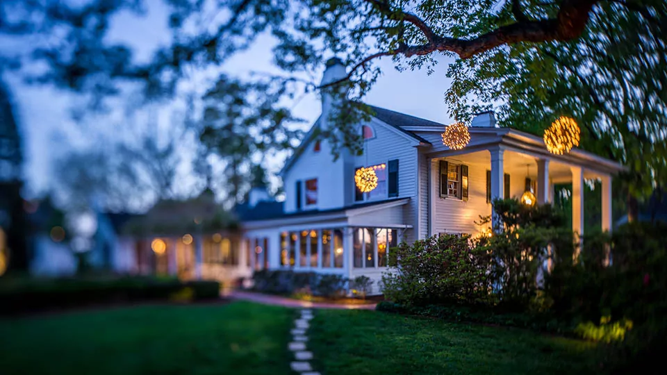 A charming two-story white house is lit by warm, glowing lights at dusk. The house features a porch adorned with hanging spherical light fixtures. The surrounding yard is lush and green with a pathway leading to the front door, framed by trees in the foreground. Fearrington Village