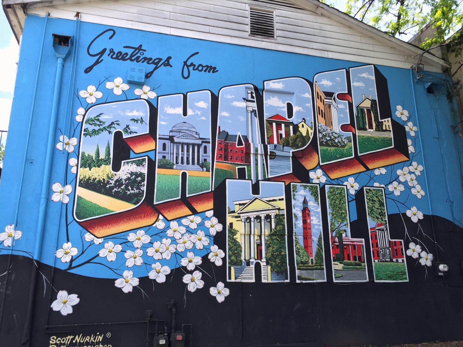 A colorful mural painted on the side of a building displays the message "Greetings from Chapel Hill." The letters in "Chapel Hill" incorporate images of local landmarks and white flowers surrounding the text. The mural is signed "Scott Nurkin" in the bottom left corner. Fearrington Village