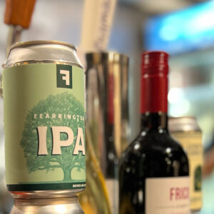 A selection of beverages including a can of IPA beer, wine bottles, and a blurred background featuring a person in a black shirt. The can of IPA is prominently visible, with other drinks and surroundings softly out of focus. Fearrington Village