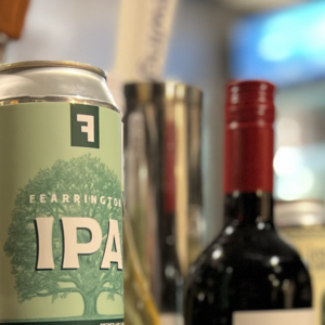 A close-up of various alcoholic beverages, including a can of IPA, a bottle of red wine, and other bottles, with a blurred background featuring a person holding a cup. Fearrington Village