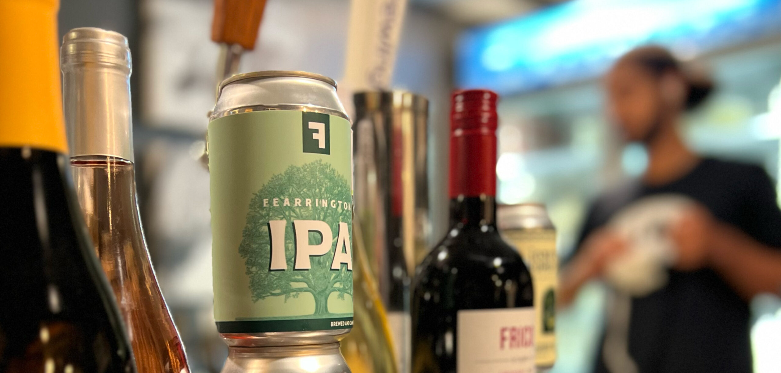 A selection of beverages including a can of IPA beer, wine bottles, and a blurred background featuring a person in a black shirt. The can of IPA is prominently visible, with other drinks and surroundings softly out of focus. Fearrington Village