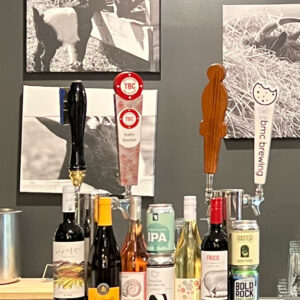 A bar counter with assorted wine bottles, beer taps, and a variety of beverages. Two black and white photos of goats are displayed on the wall behind the counter. The beer taps include labels for TBC, BBC Brewing, and other brands. Fearrington Village