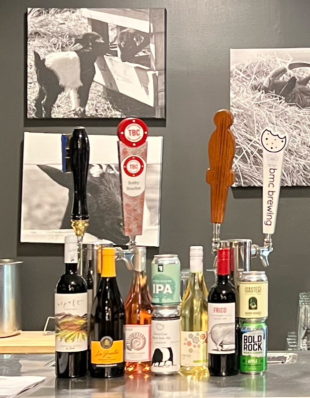 A bar counter with assorted wine bottles, beer taps, and a variety of beverages. Two black and white photos of goats are displayed on the wall behind the counter. The beer taps include labels for TBC, BBC Brewing, and other brands. Fearrington Village