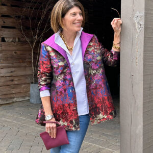 A woman stands outdoors leaning against a wooden post. She is wearing a colorful jacket with shades of red, pink, and purple, over a white shirt, and blue jeans. She holds a purple clutch and has short brown hair. Greenery and a wooden fence are in the background. Fearrington Village